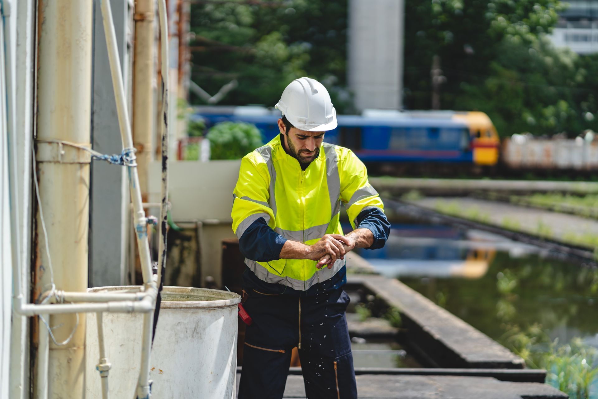 Person in high-visibility jacket and hard hat, checking wrist, near industrial equipment and train tracks.