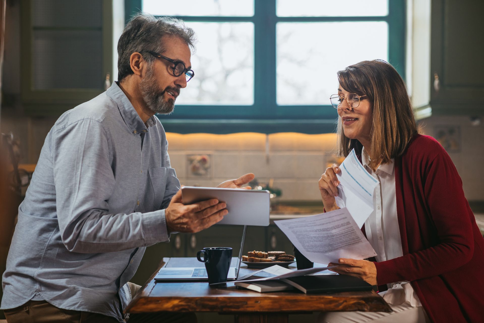 Man and woman review documents at a table; man holds tablet, woman holds papers.