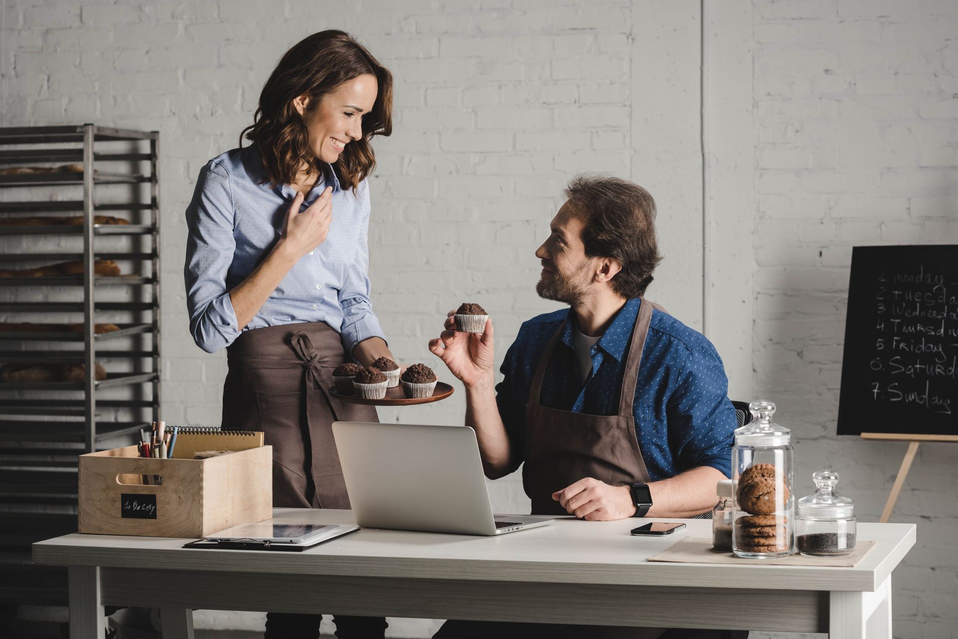 Man offers woman a muffin at a bakery table; both smile. Aprons, muffins, laptop, and chalkboard are present.