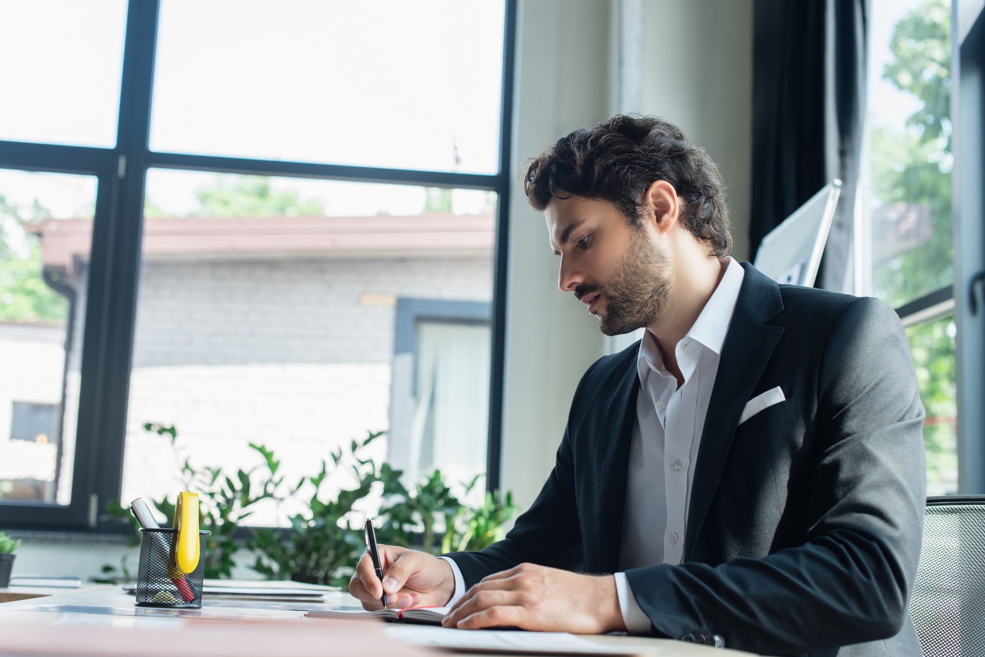 Man in suit writing at desk near a window, with plants in view.