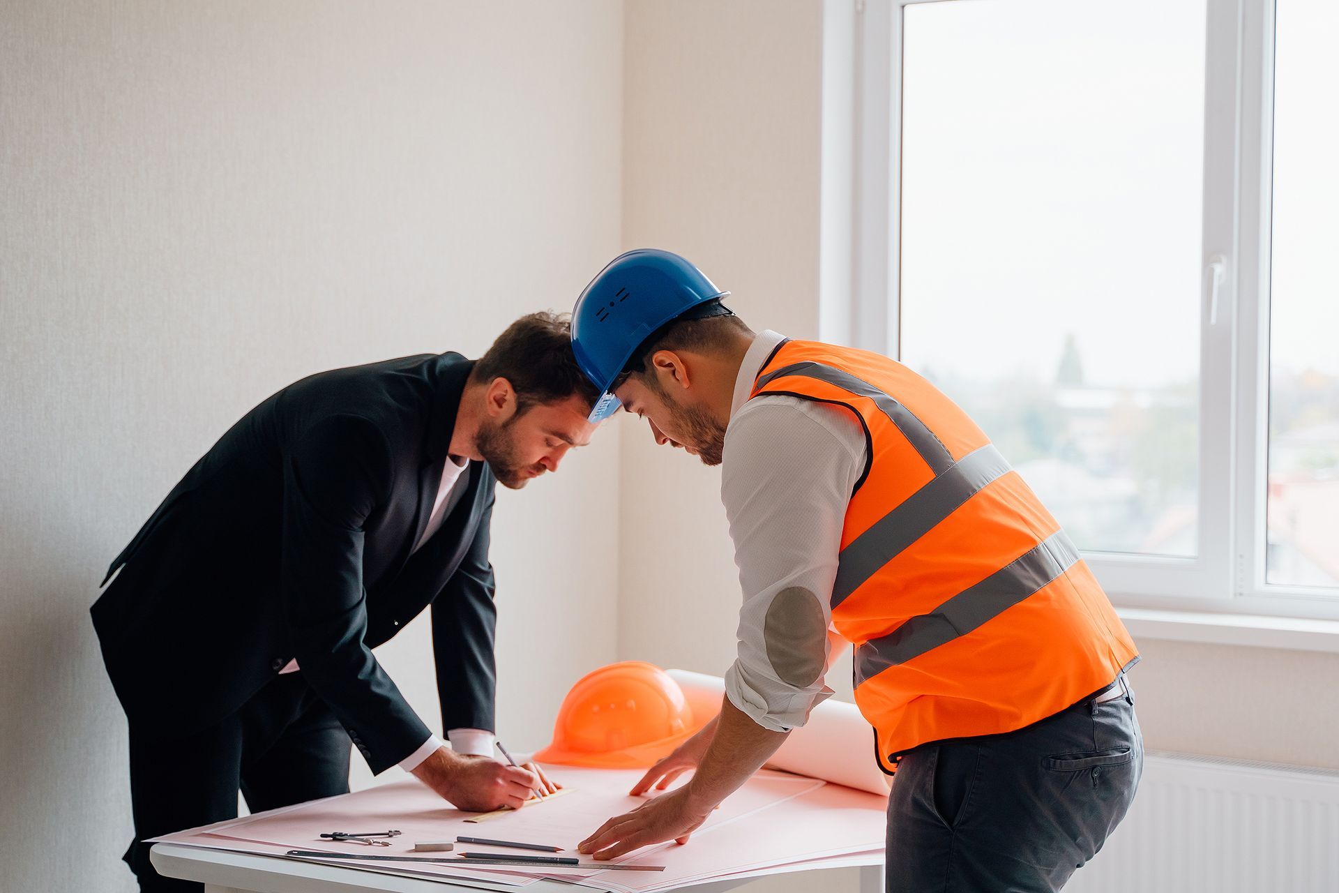 Two men, one in suit and one in work gear, review blueprints at a table, orange hard hat and vest present.