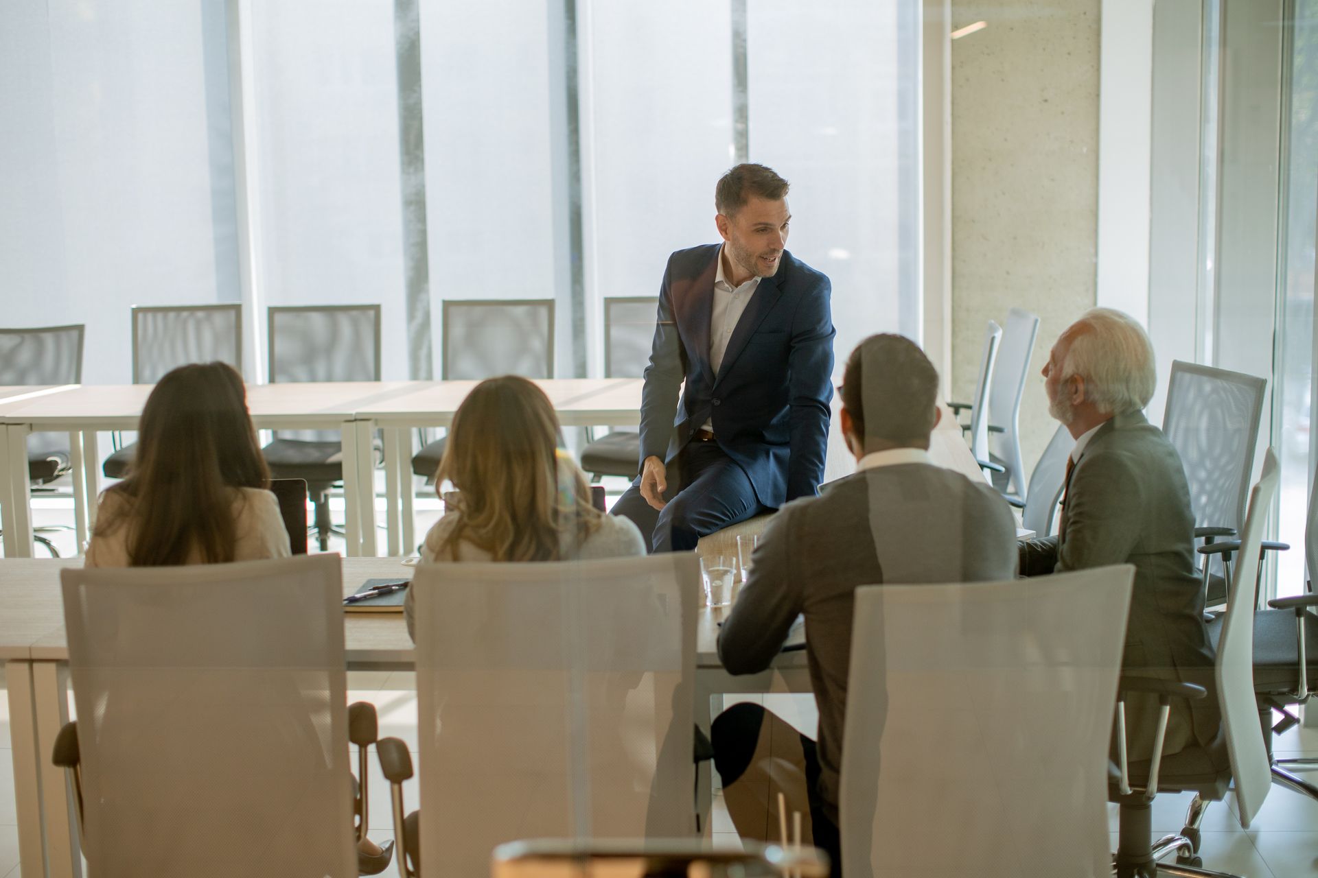 Business meeting in a bright room. A man stands, speaking to four seated colleagues around a table.