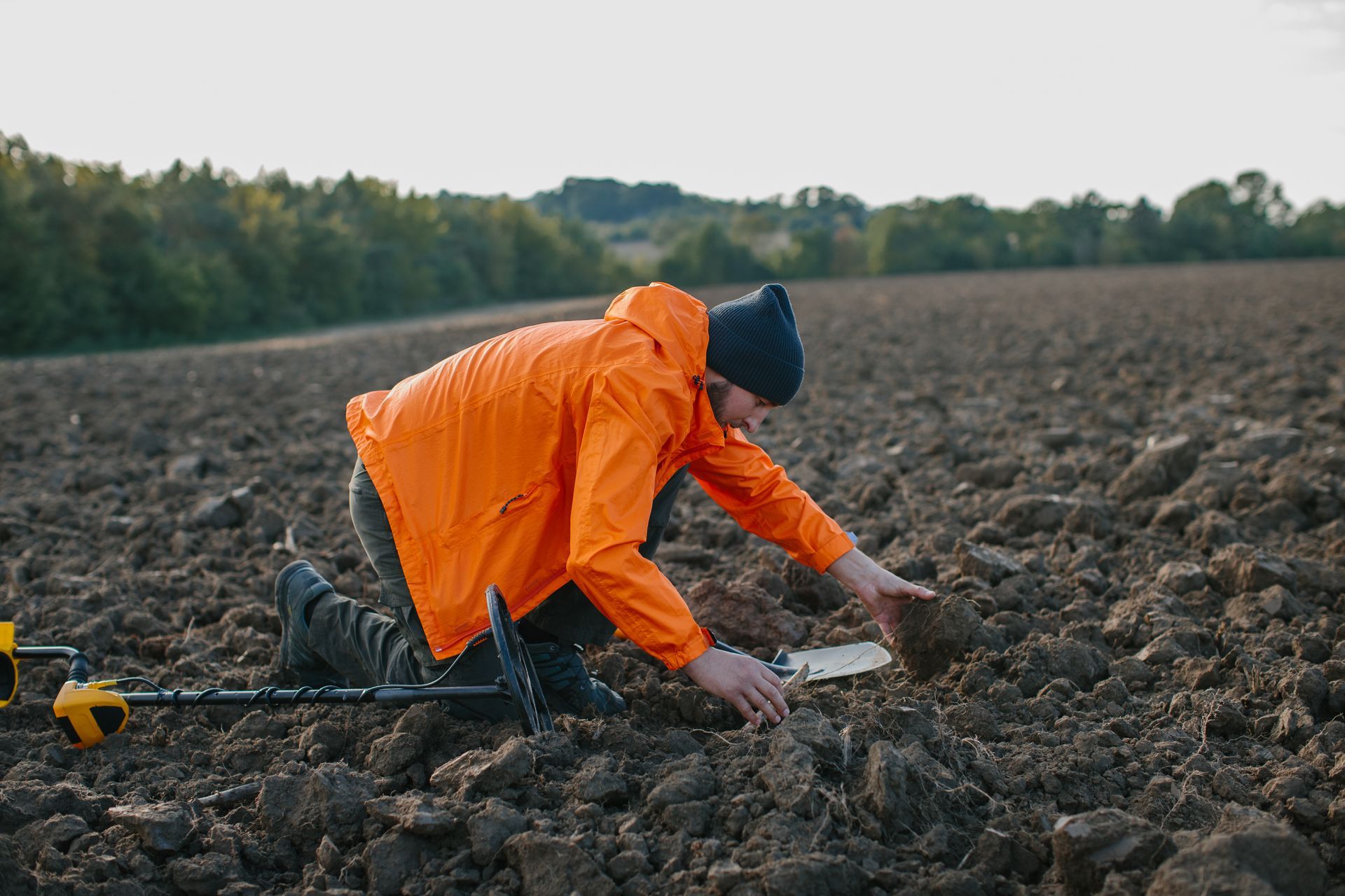 Person kneeling in a plowed field, wearing orange jacket and a black hat, metal detecting.