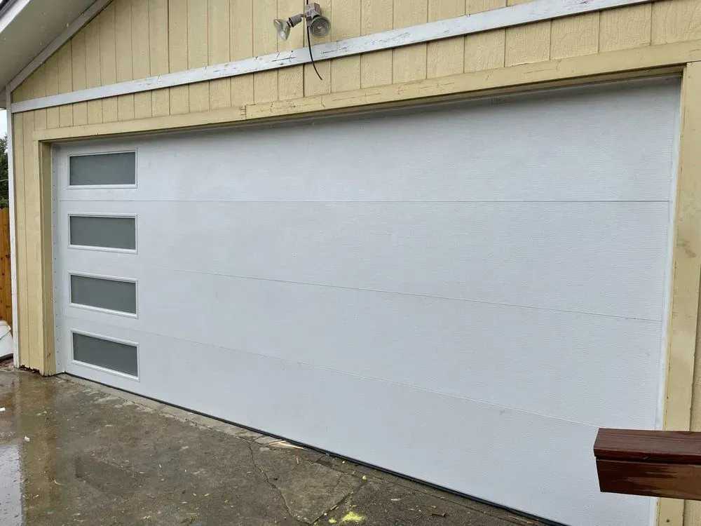White garage door with four frosted windows beside a wet driveway and beige siding