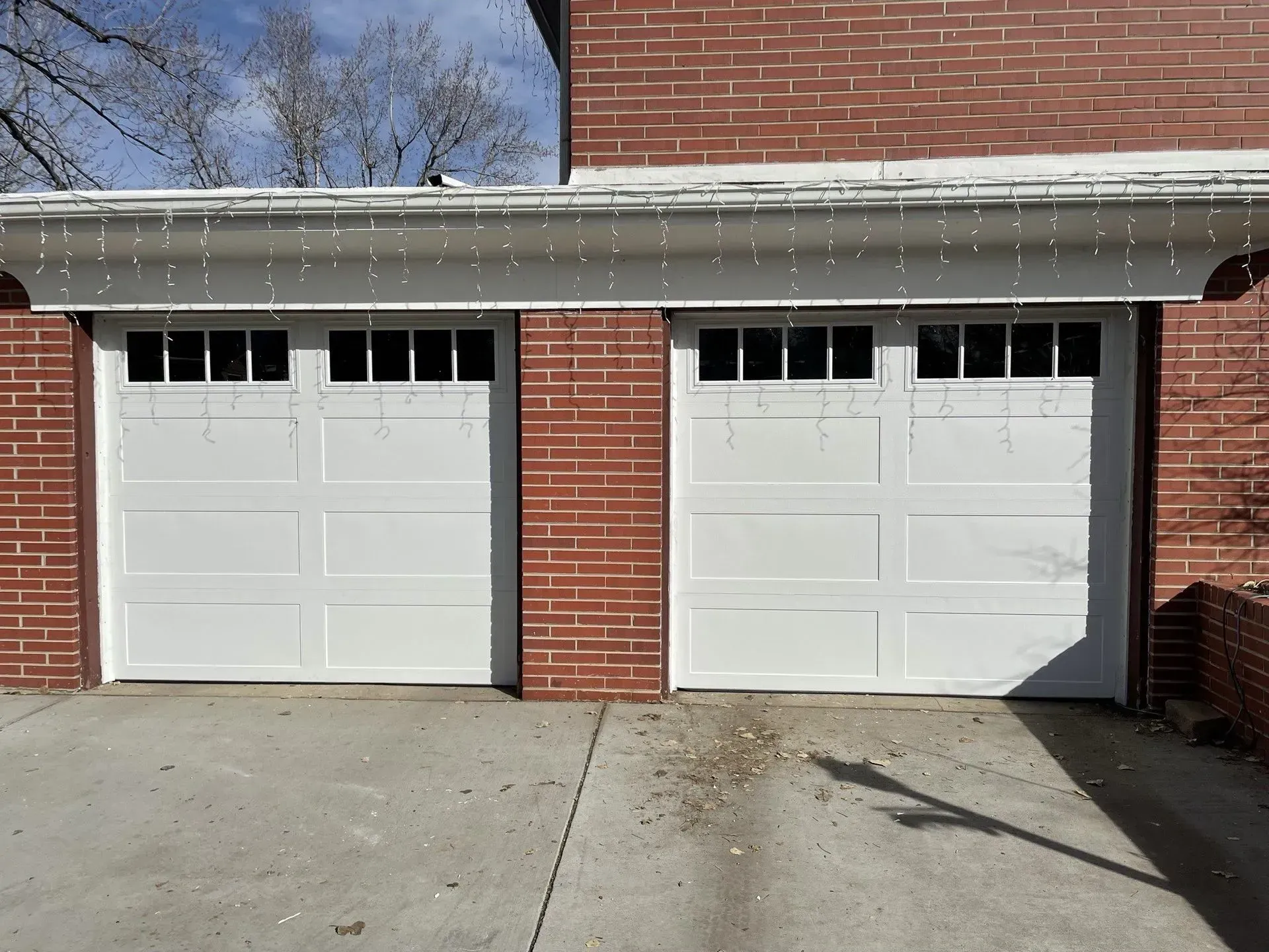Two white garage doors on a brick house with a concrete driveway and snow along the roofline.