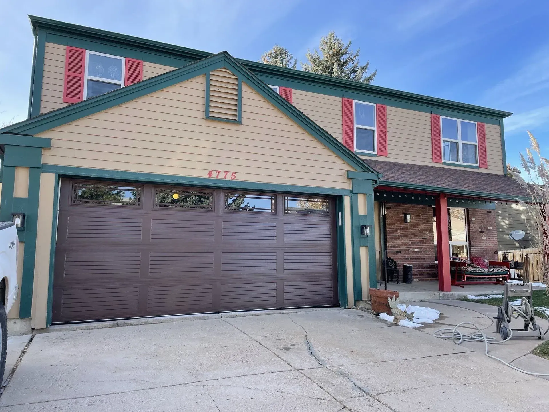 Two-story beige house with teal trim, red shutters, and a brown garage door on a snowy driveway