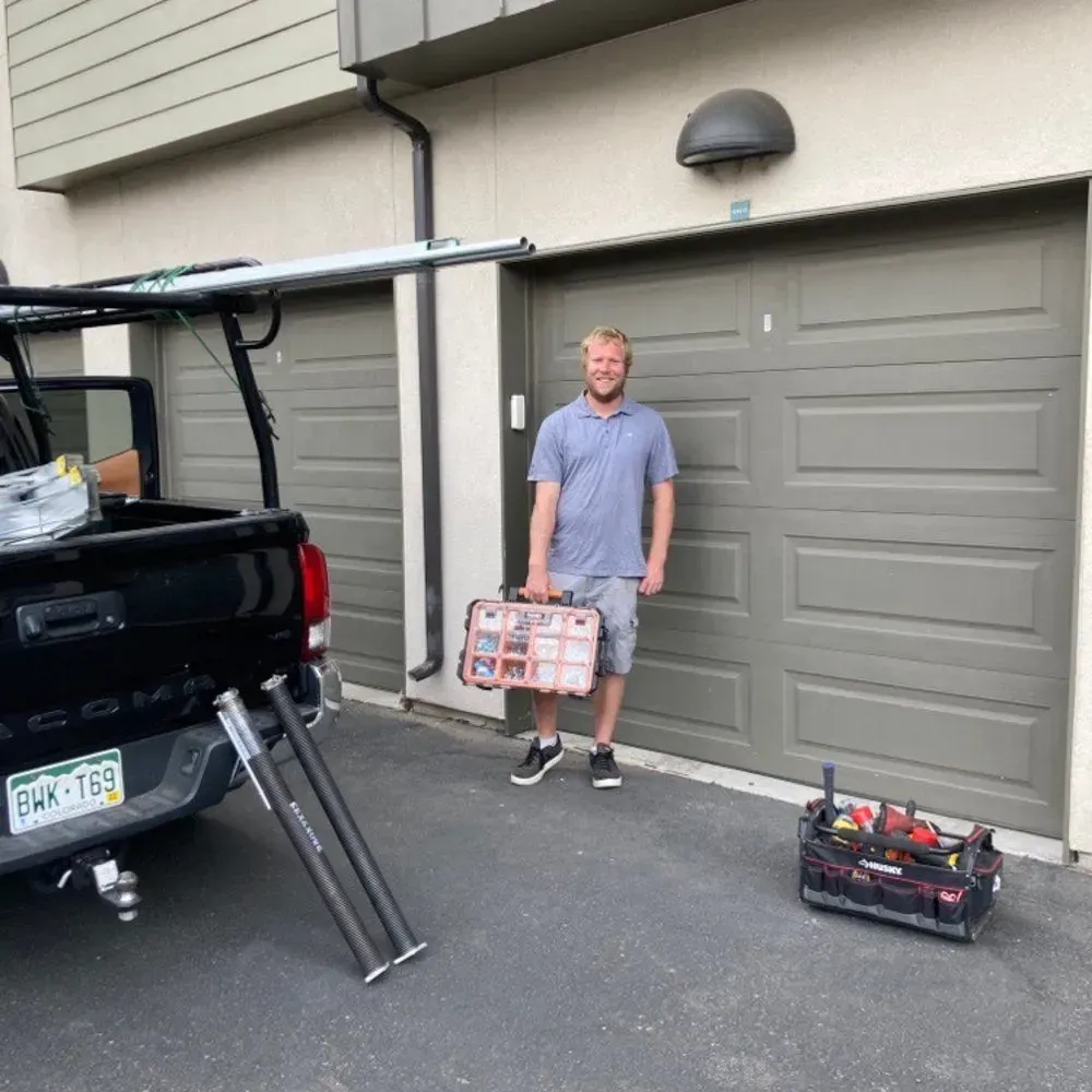 Man standing beside an open truck bed, holding a toolbox in a driveway outside a garage