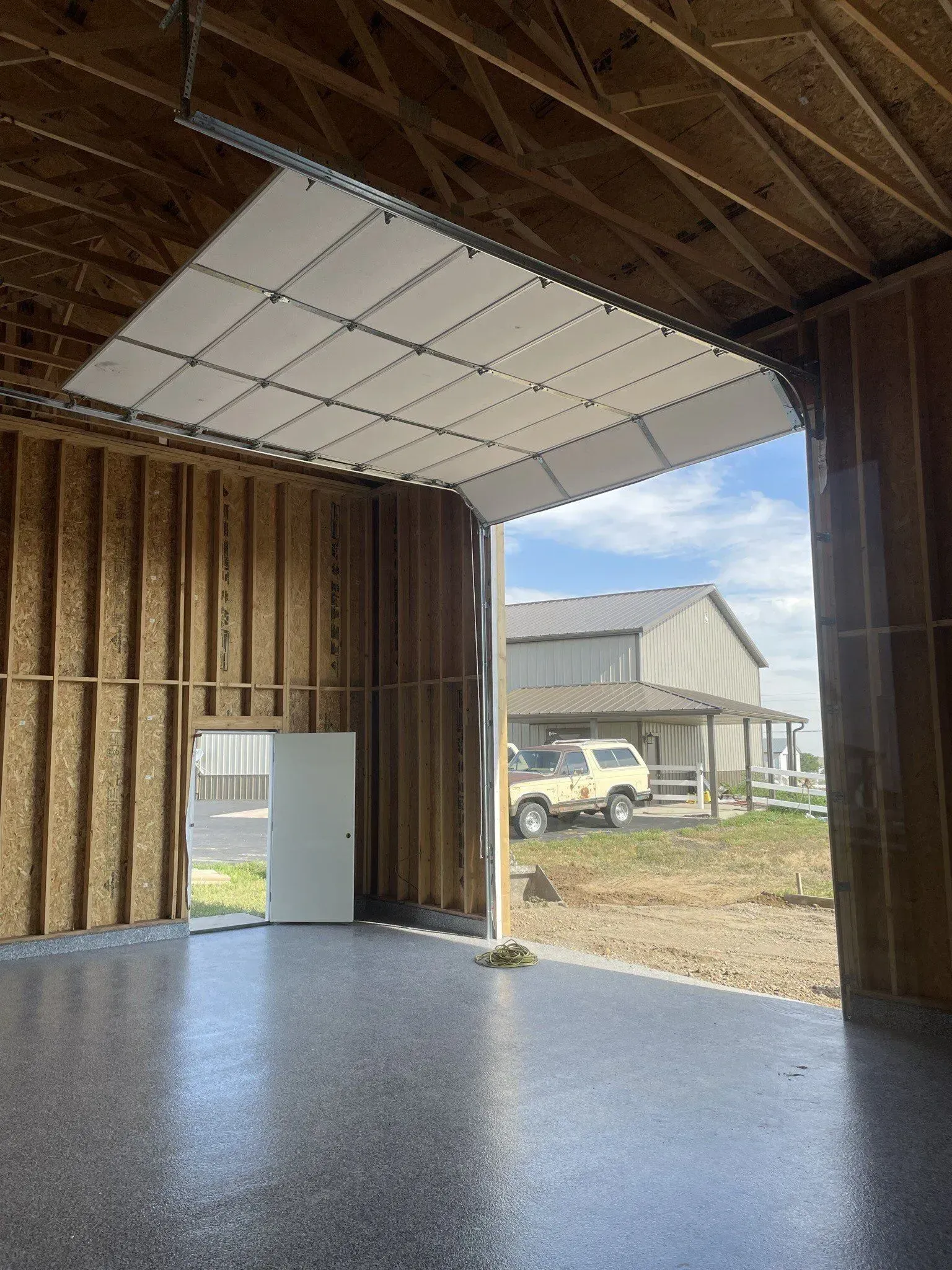 Empty unfinished garage with exposed wood framing, white ceiling panels, and a view of buildings outside.