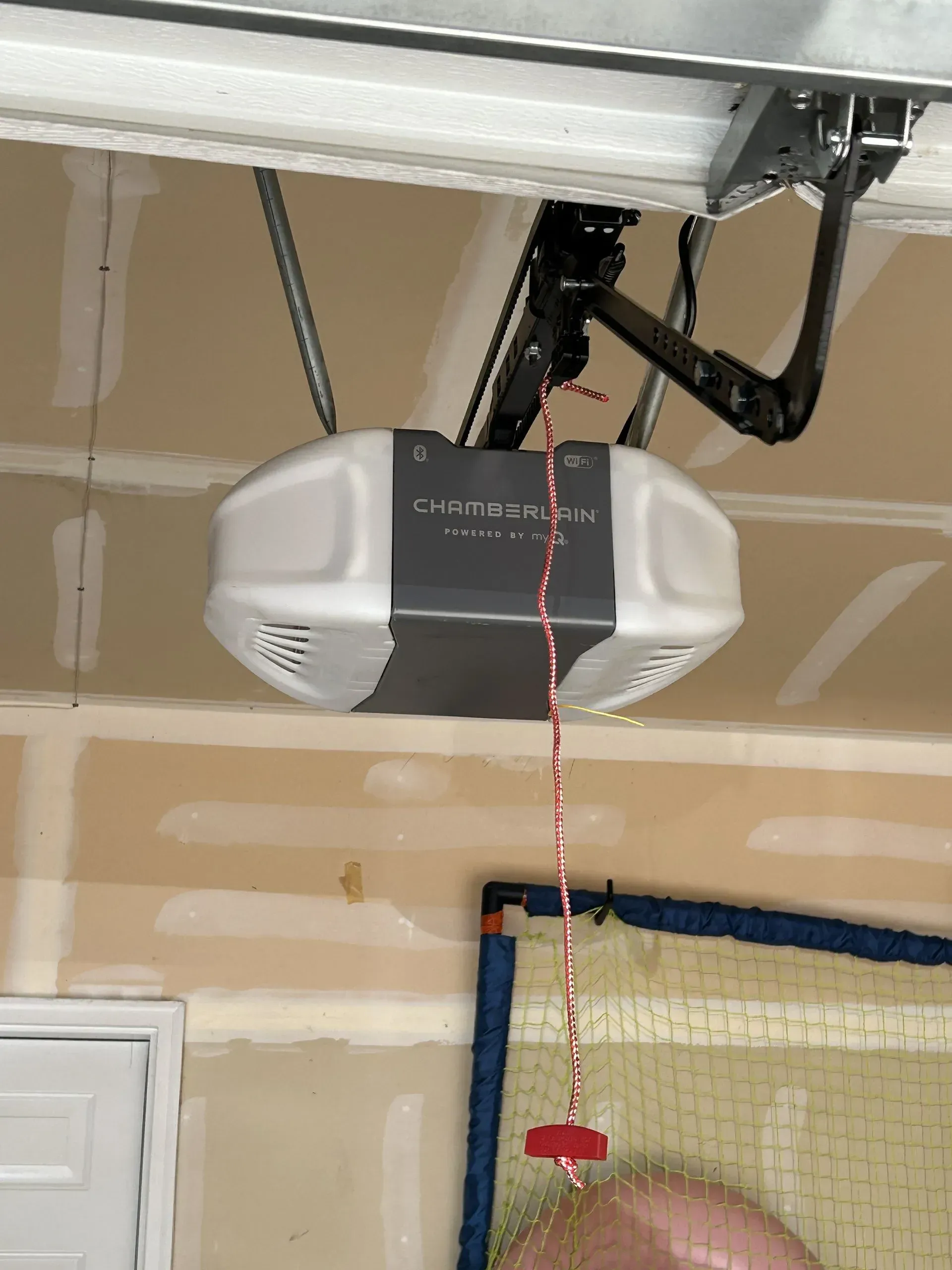 Garage door opener mounted on ceiling with red pull cord and white garage door below