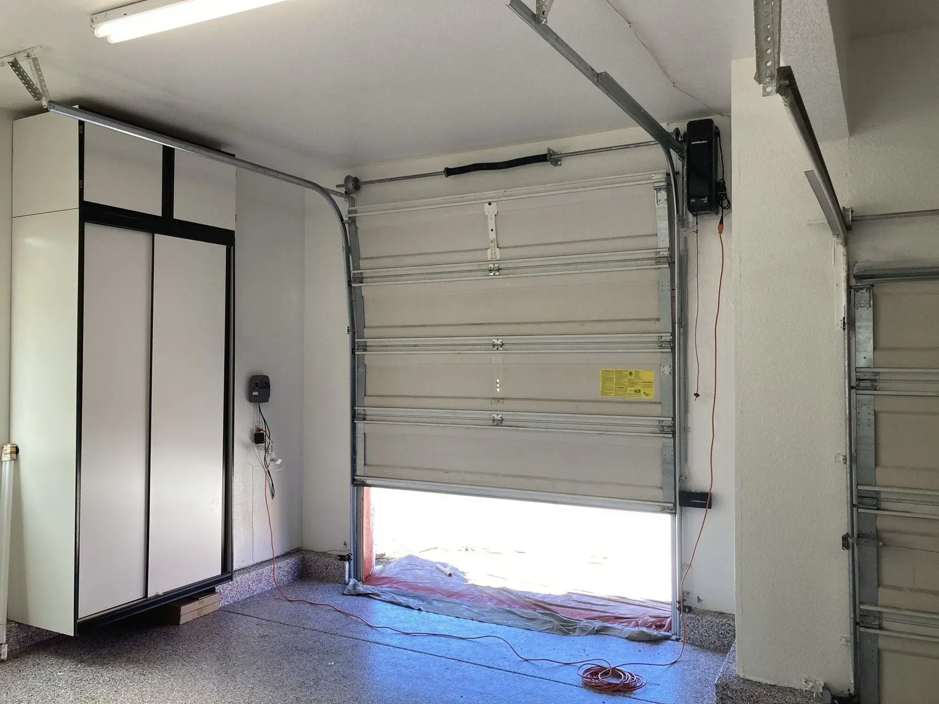 Interior of a garage with a partially open overhead door and white cabinets on the left.