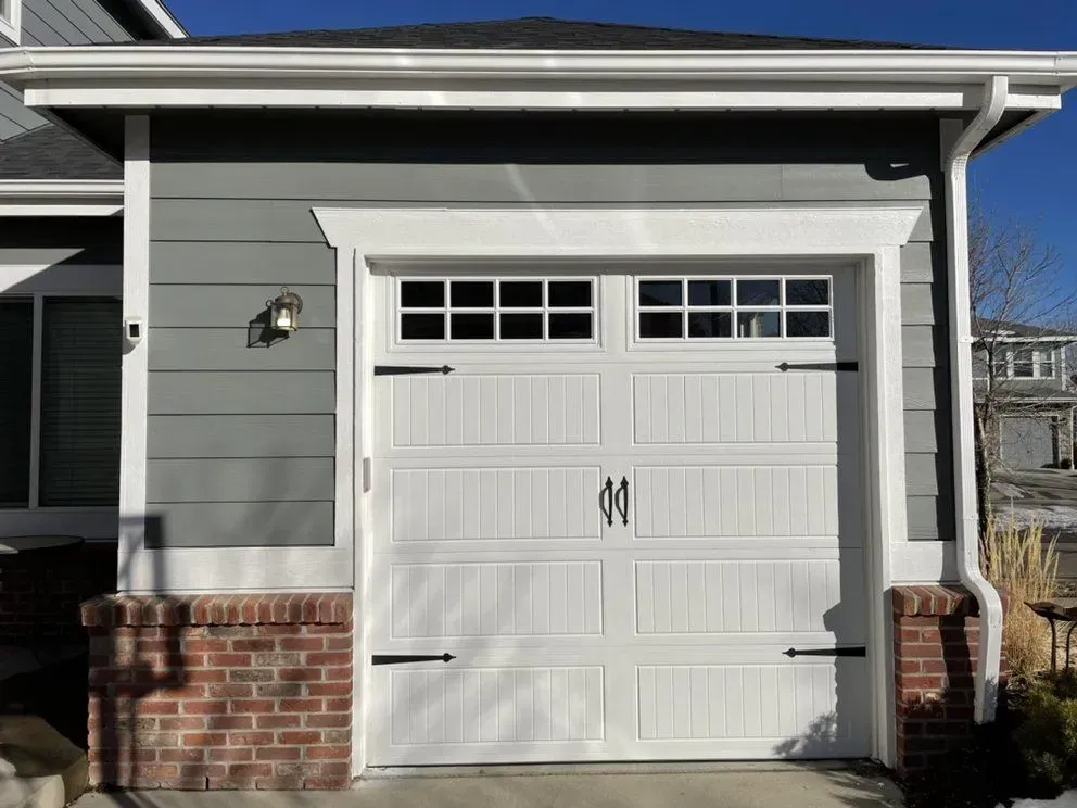 White garage door on a gray house with brick accents and a wall light under a blue sky
