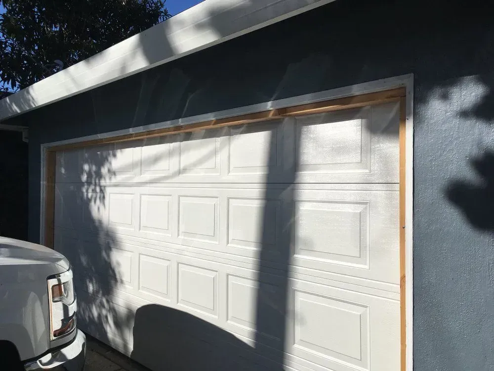 White garage door on a blue house, partly shaded by trees, with a white vehicle parked at left.