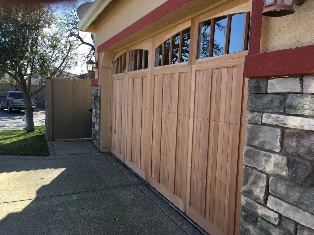 Wooden garage door beside a stone wall and driveway, with upper windows and a side gate.
