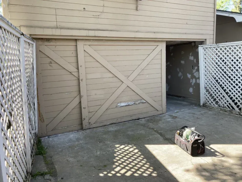 Backyard shed with closed beige double doors, white lattice fencing, and a duffel bag on the concrete patio.