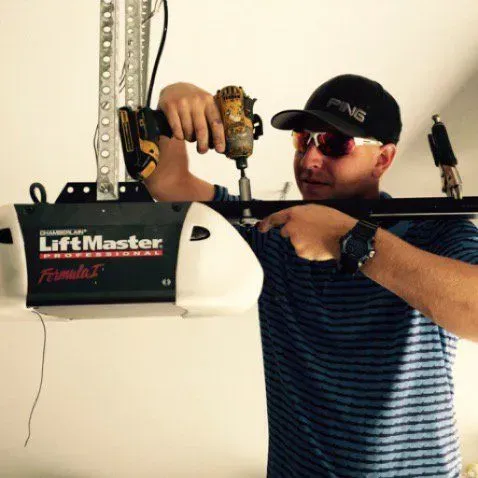 Technician installing a LiftMaster garage door opener with a drill, wearing a cap and sunglasses.