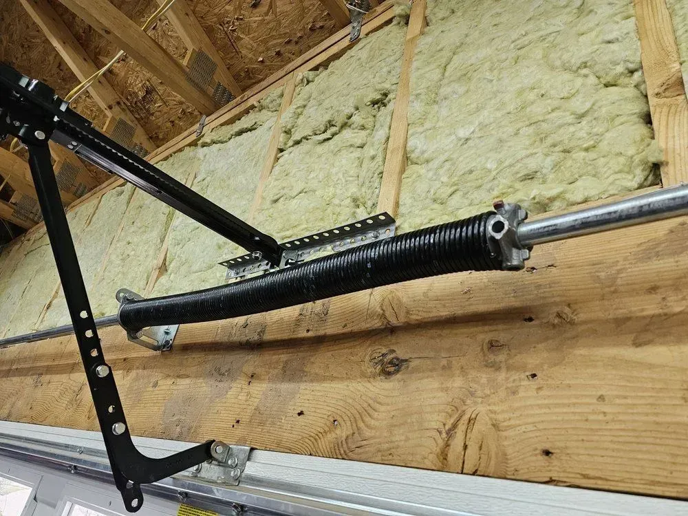 Black garage door torsion spring and track mounted on wooden beam in unfinished attic area