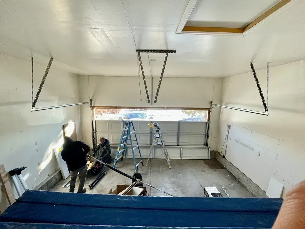 Garage interior under renovation with ladders, tools, and workers near an open garage door