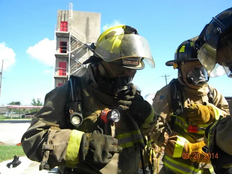 Two firefighters are standing next to each other in front of a fire training tower on a sunny day