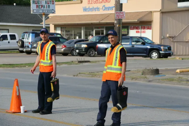 Two men wearing orange vests and safety vests are standing in front of a building holding fire boots for MDA fill the boot.