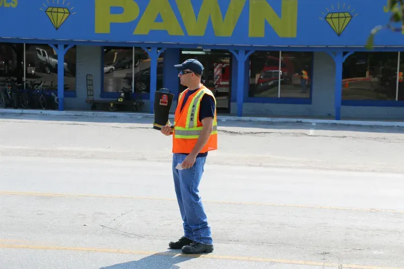 A man in an orange vest is standing in front of a pawn shop holding a fire boot