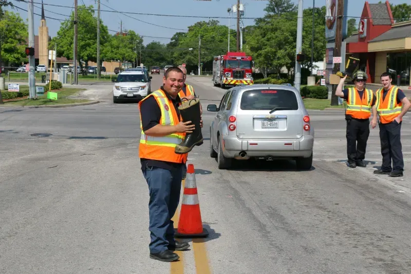 A man in a safety vest is standing next to a traffic cone holding a fire boot 