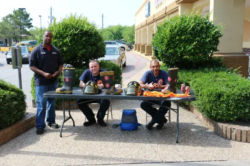 Three men are sitting at a table with fire helmets and boots on it.