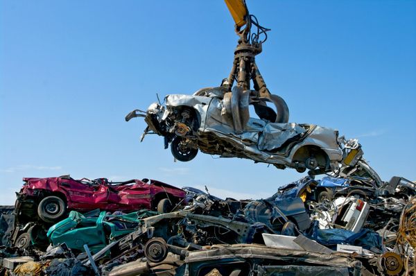 A crane lifts a crushed car at a junkyard, blue sky in the background.