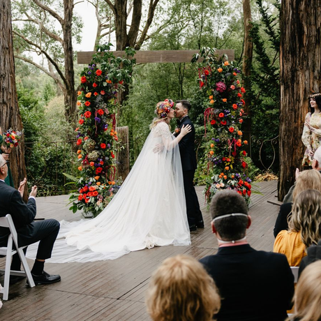 Bride and groom kiss under a flower-covered arch at an outdoor wedding ceremony
