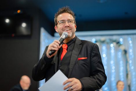 Man speaking into a microphone at an event, wearing a dark suit with a red tie and pocket square, holding papers