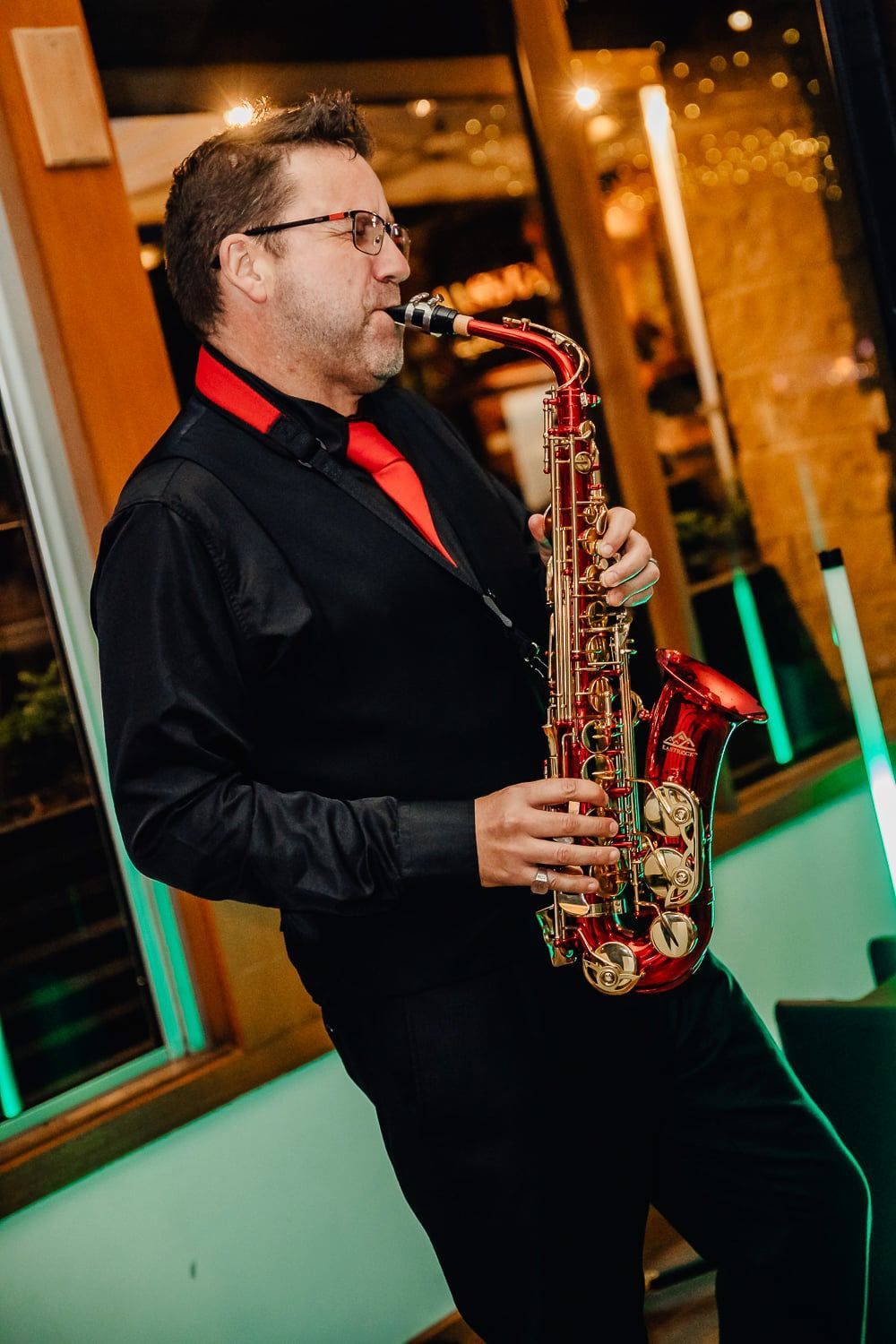 Man playing a gold saxophone in a black shirt and red tie at a warmly lit indoor venue