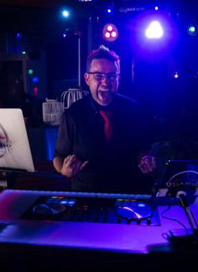 Person at a DJ booth under purple-blue stage lights, holding a microphone and smiling.