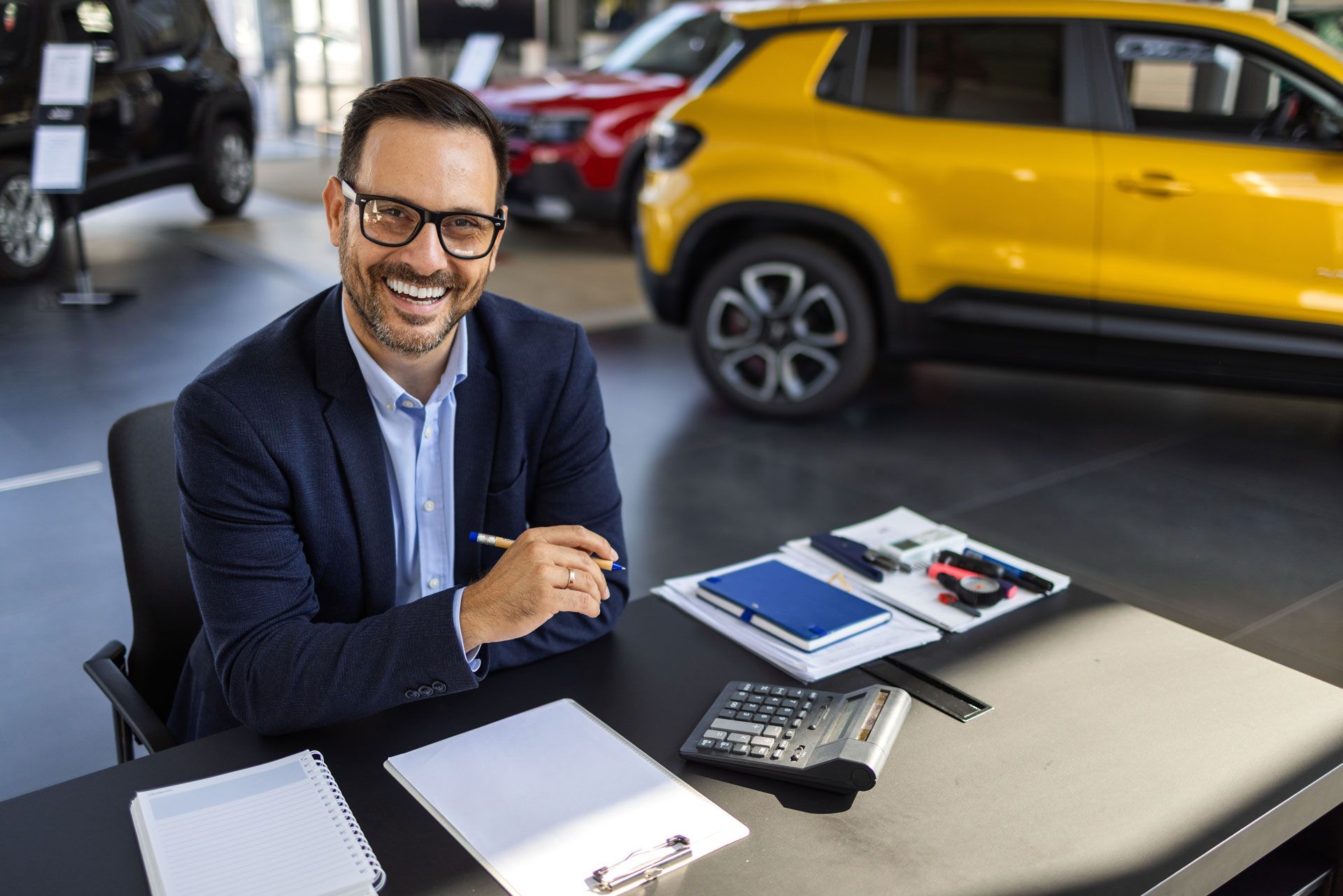 Car salesman smiling behind a desk, showroom background, yellow car visible.