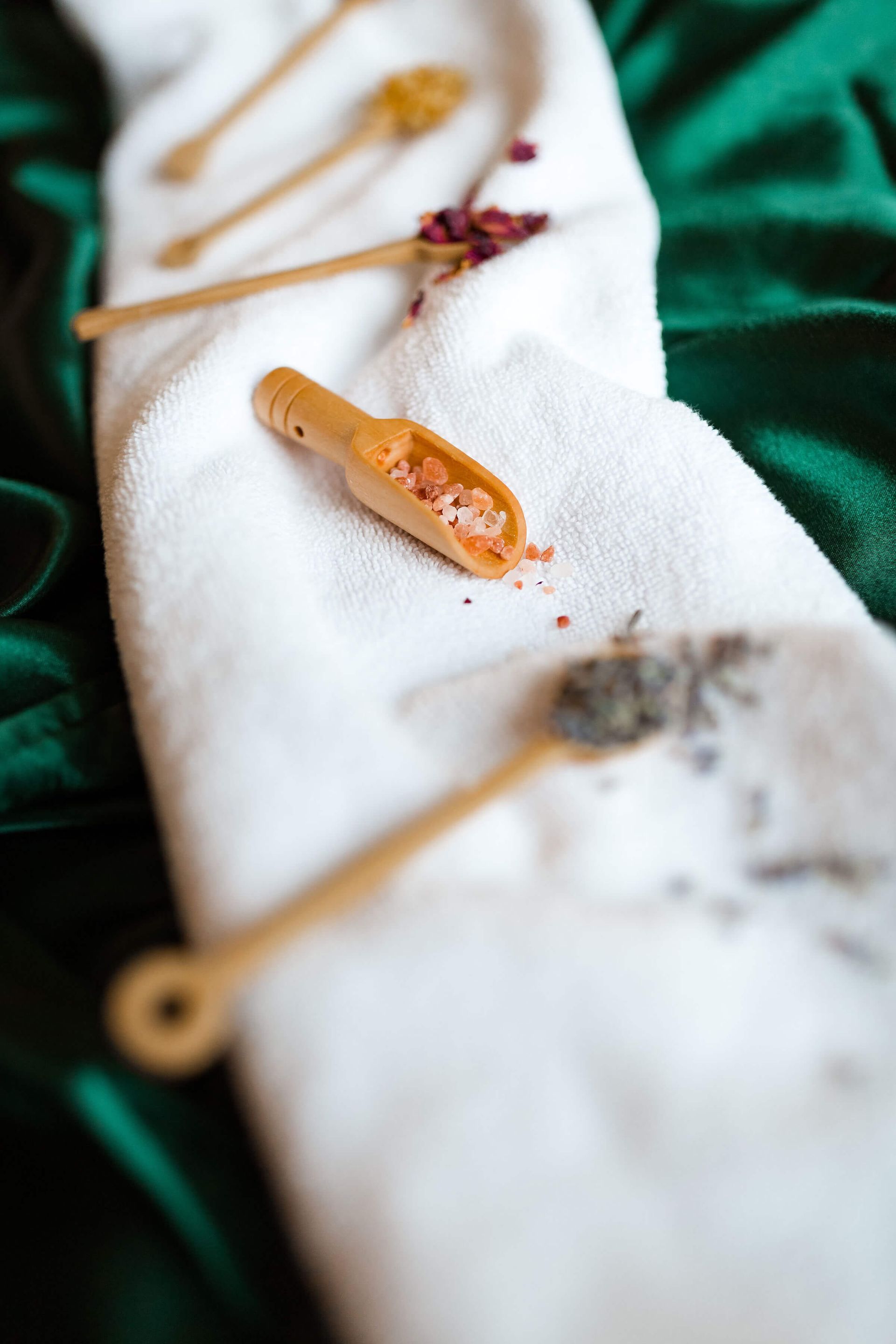 A row of wooden spoons filled with different types of bath salts and herbs on a white towel.