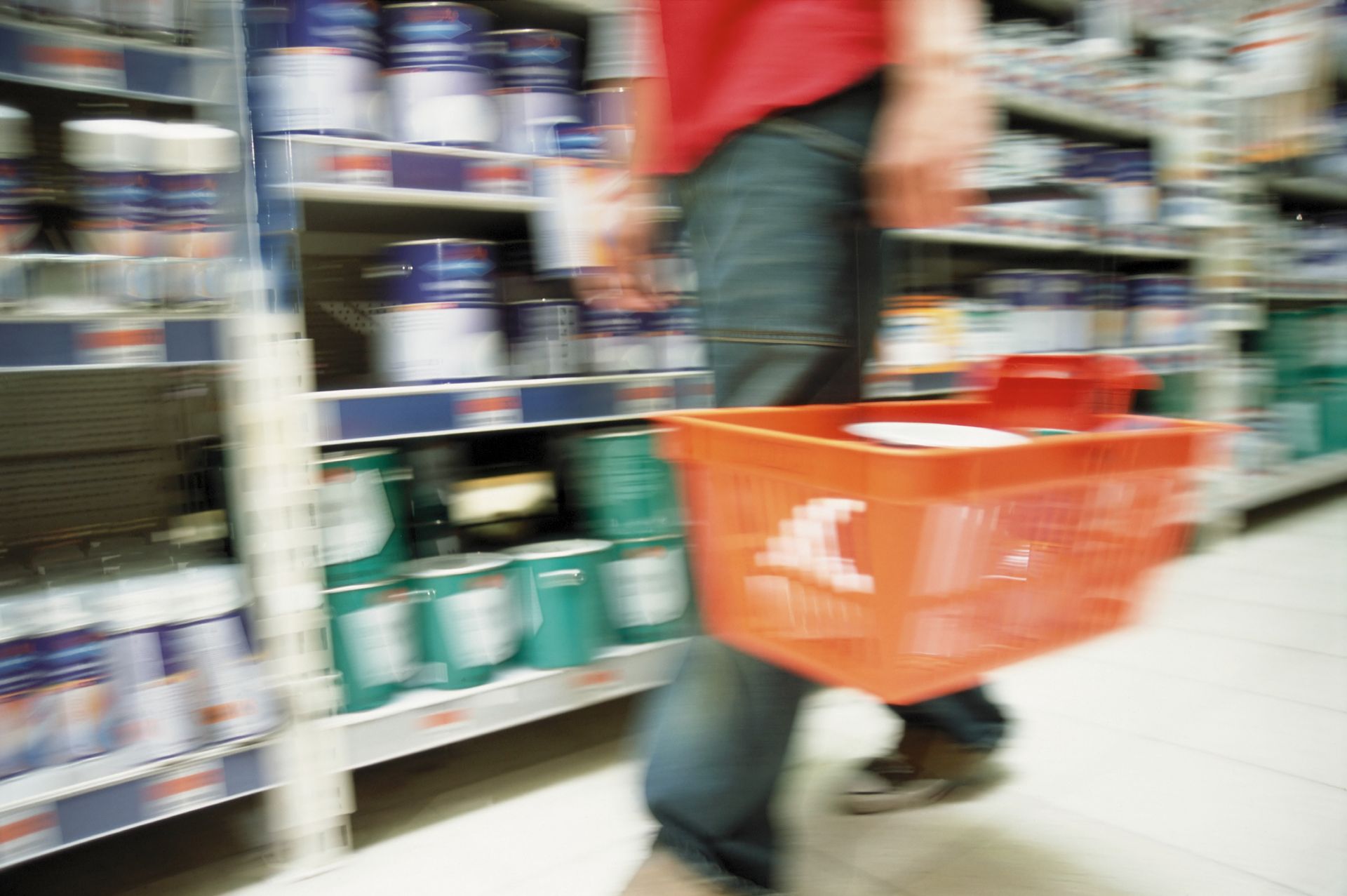 Person with an orange shopping basket browsing paint shelves in a store.