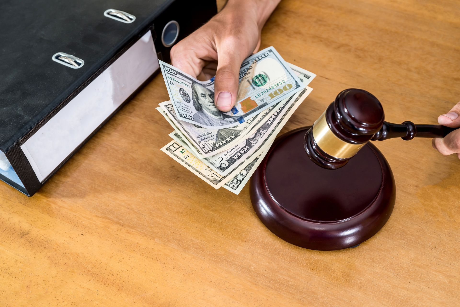 Hand holding cash next to a gavel and a binder on a wooden surface.