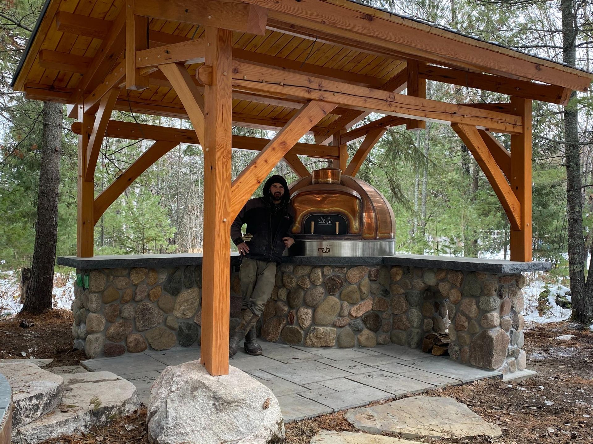 A man is standing under a wooden structure next to a pizza oven.