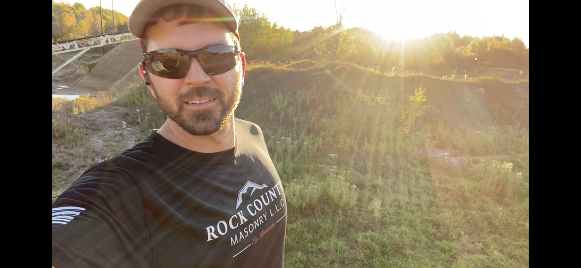 A man wearing sunglasses and a hat is taking a selfie in a field.