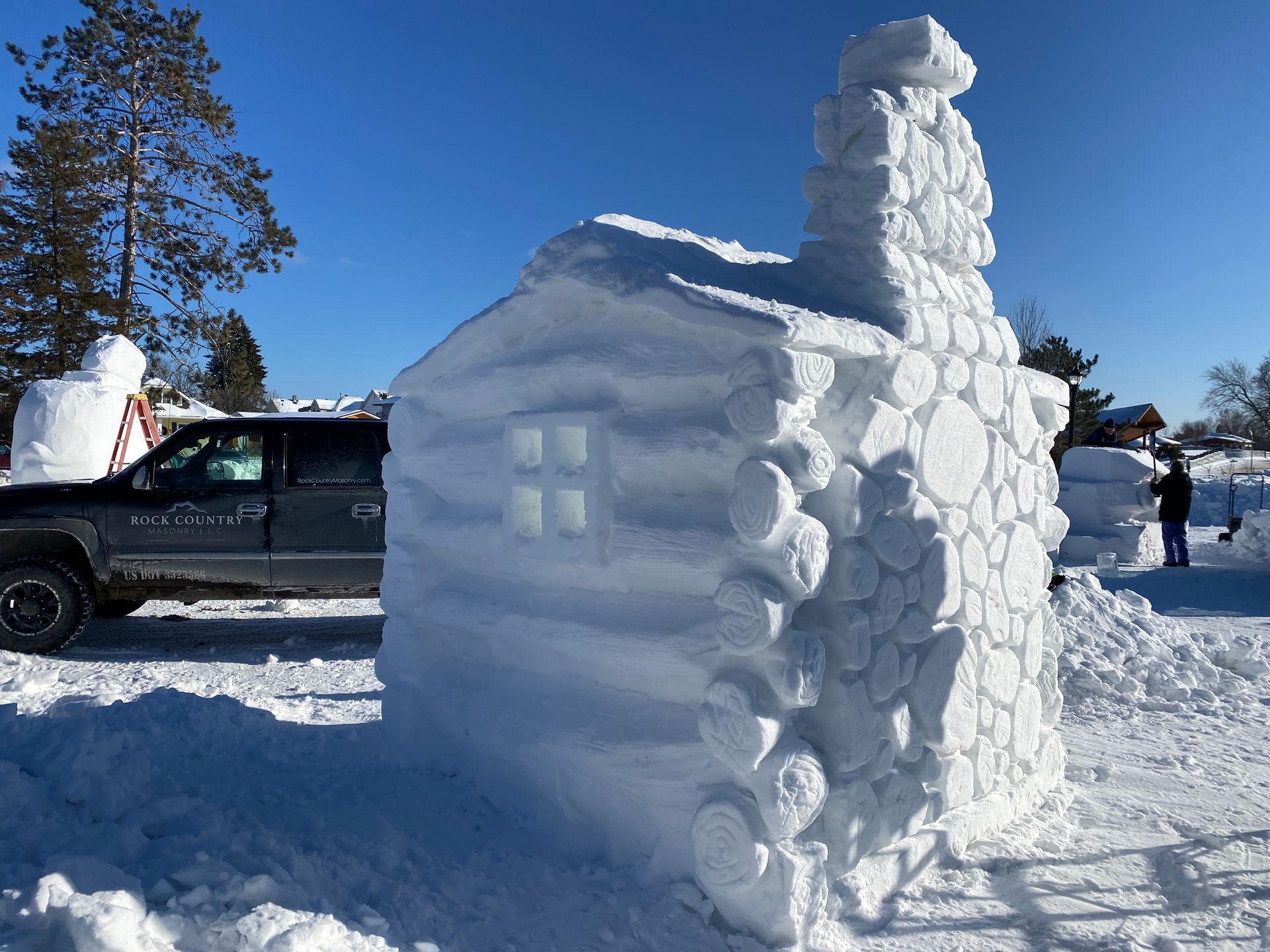 A car is parked next to a snow sculpture of a log cabin.