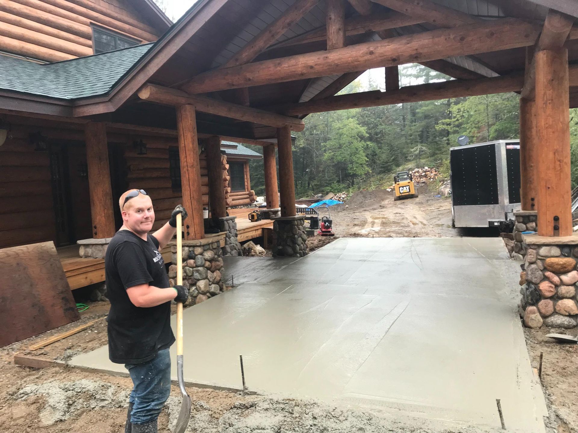 A man is standing in front of a log cabin holding a shovel.