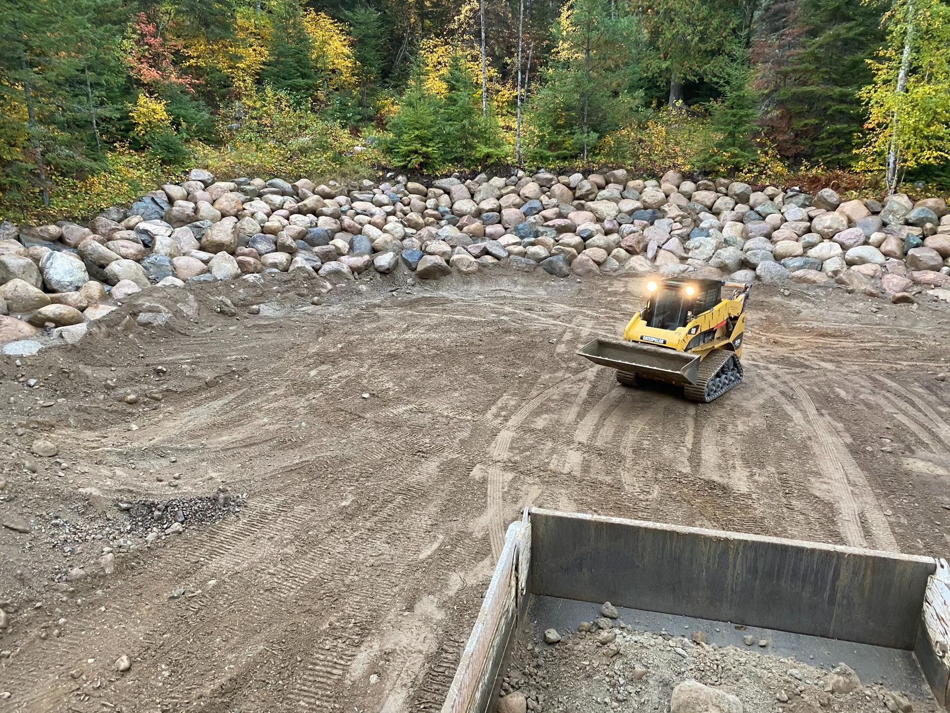 A bulldozer is driving down a dirt road next to a dumpster filled with rocks.