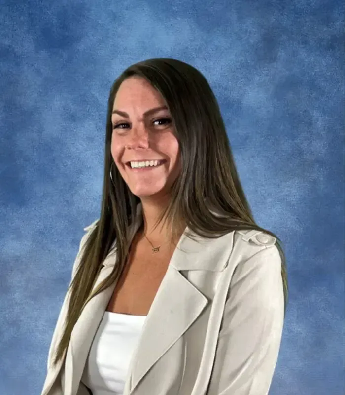 Woman with long brown hair smiles, wearing a white top and light beige blazer, against a blue backdrop.