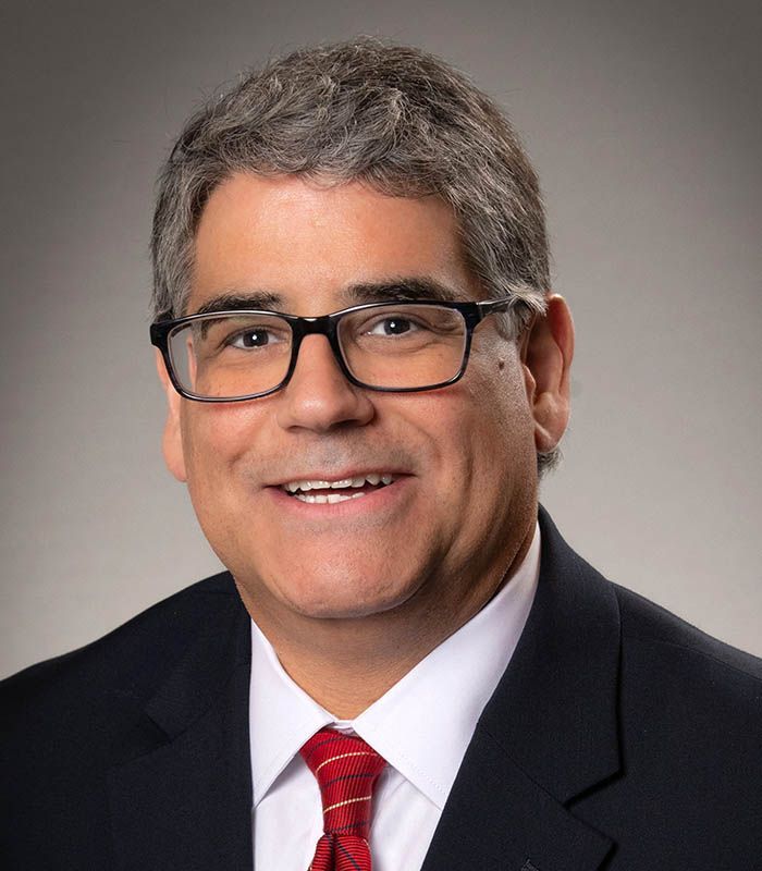 Man with glasses and a red tie smiles, likely a professional headshot.