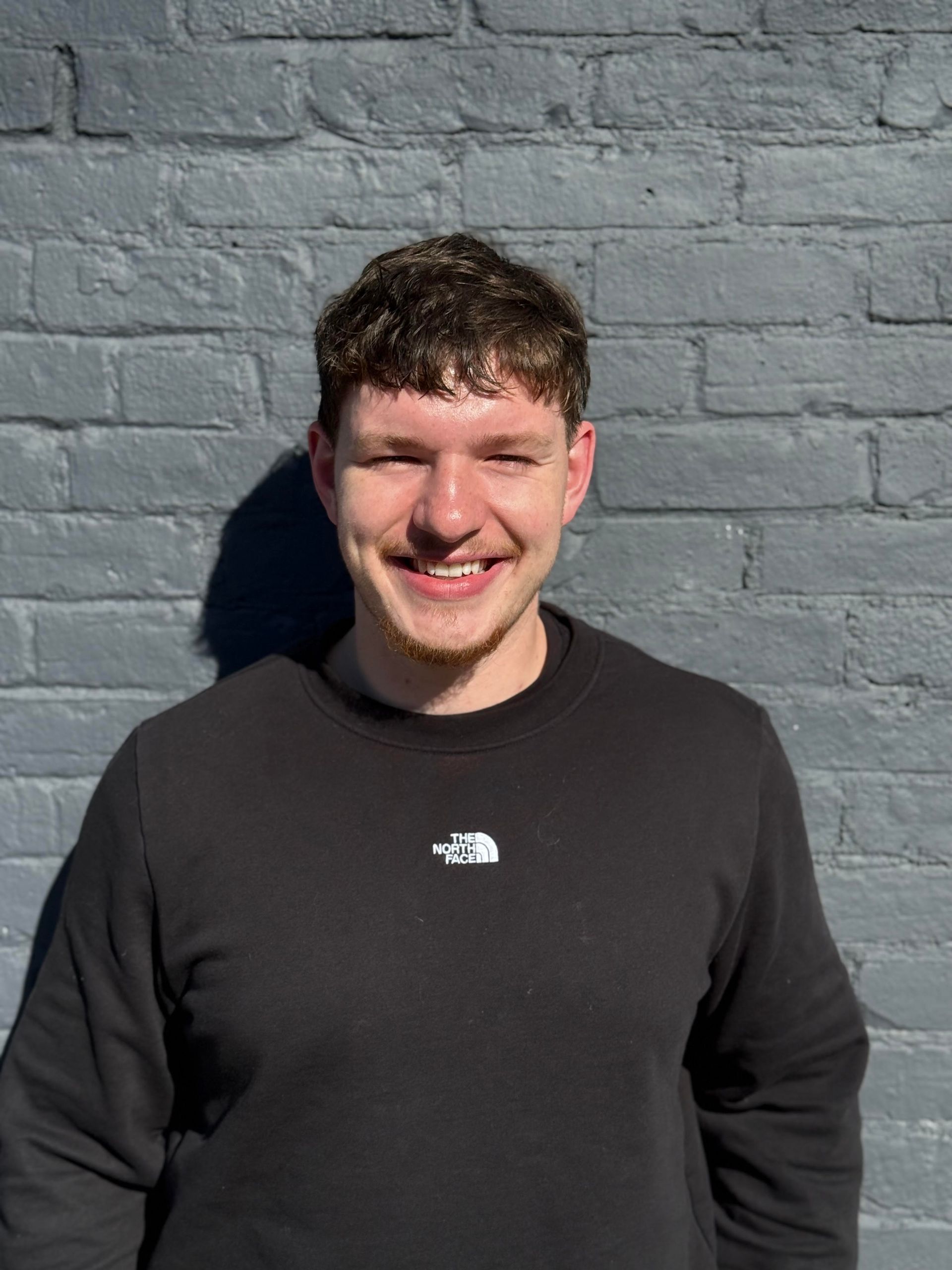 Man smiling, wearing a black sweatshirt with a white logo, standing in front of a gray brick wall.