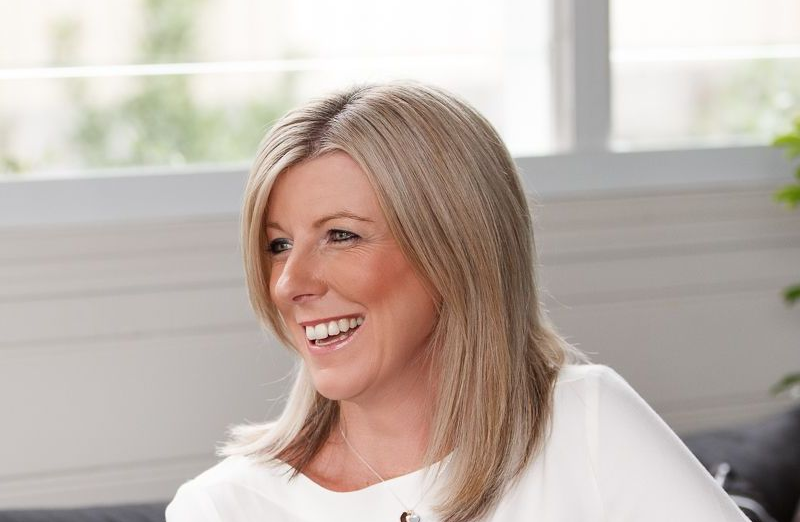 Woman with blonde hair smiles while working on a laptop. Sitting at a table indoors.