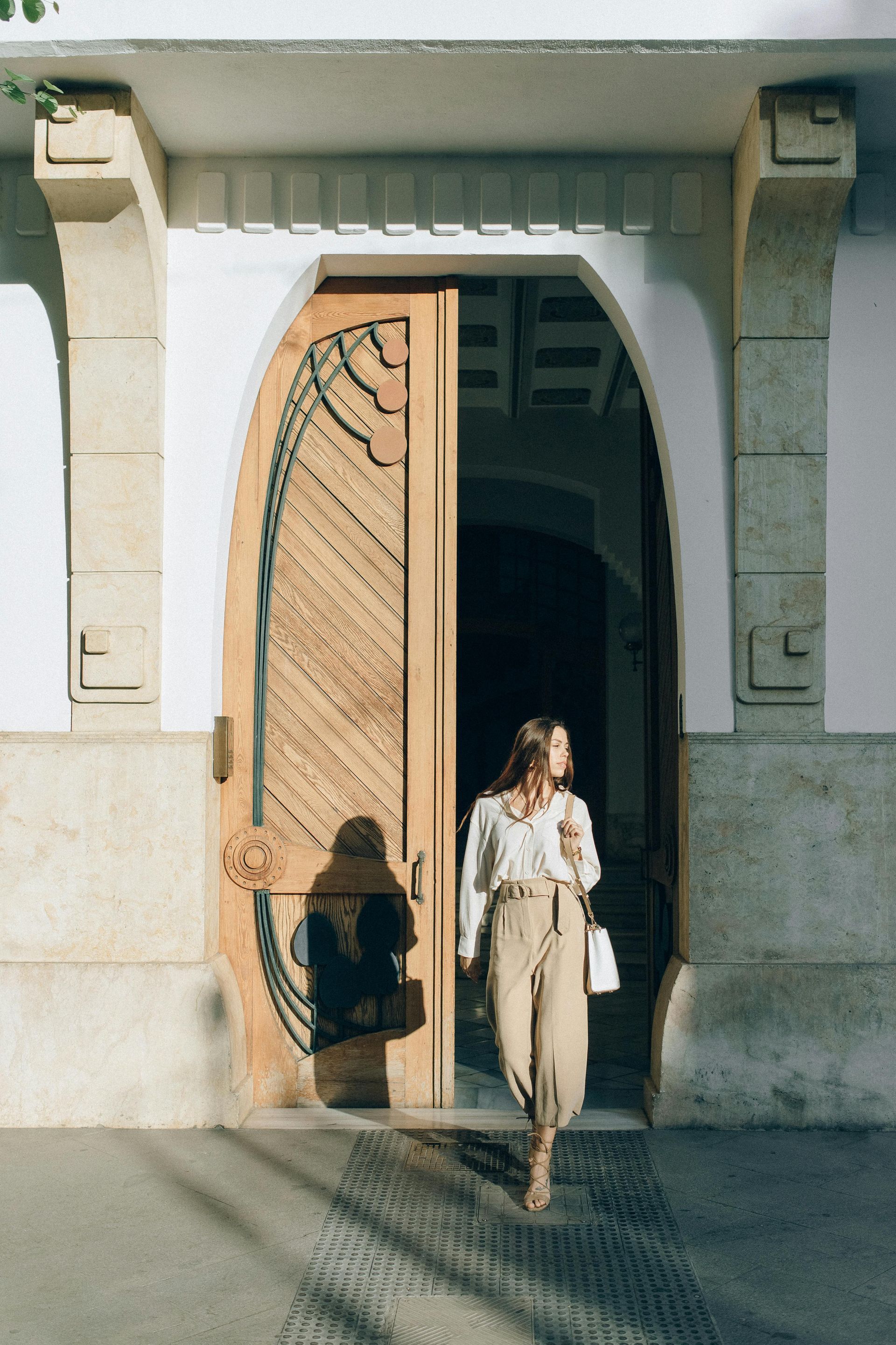 Woman exiting an ornate wooden door; sunlit exterior, wearing cream blouse, tan pants, and carrying a white bag.