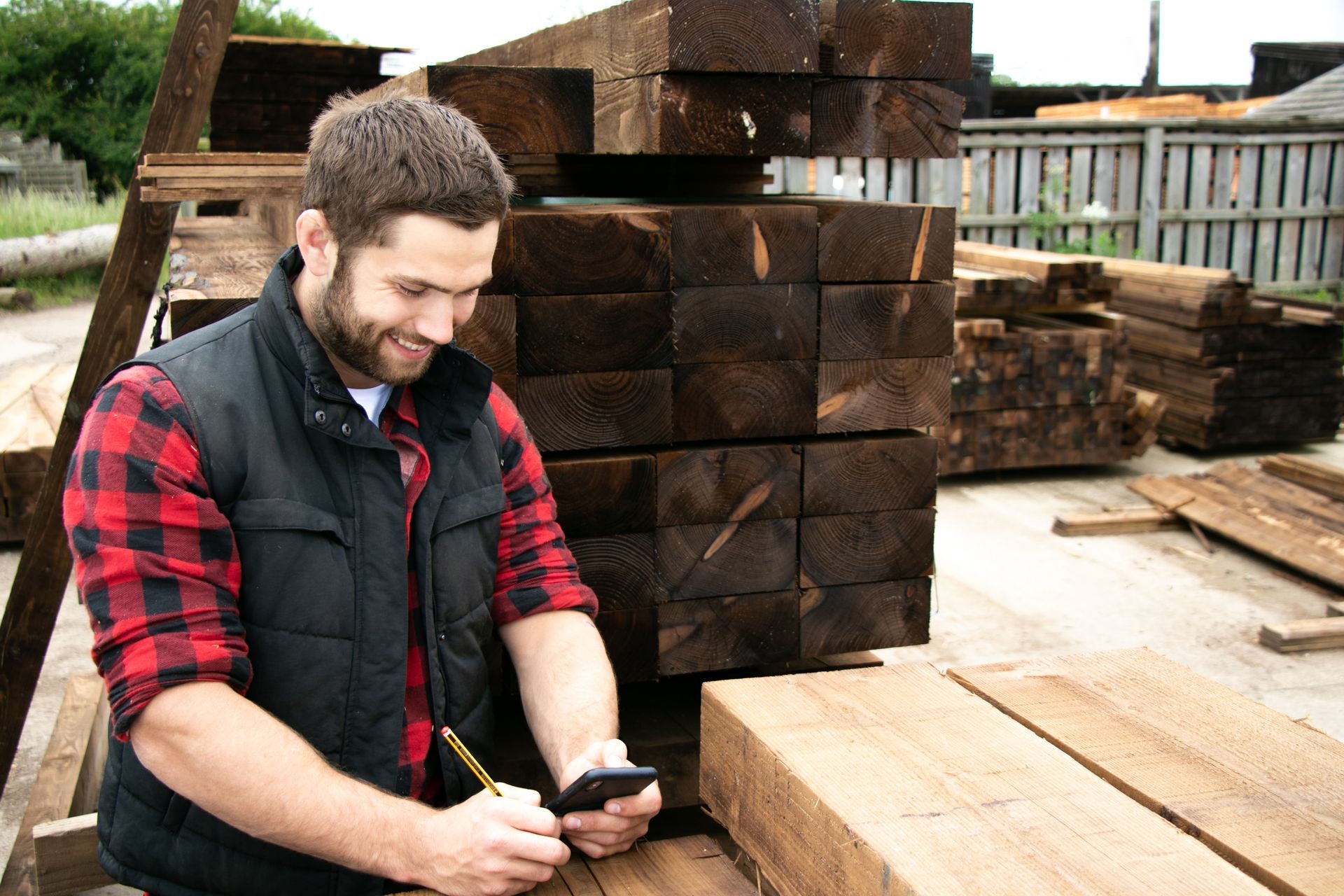 A person in a red plaid shirt and black vest smiles while holding a phone and a pencil in a lumber yard.