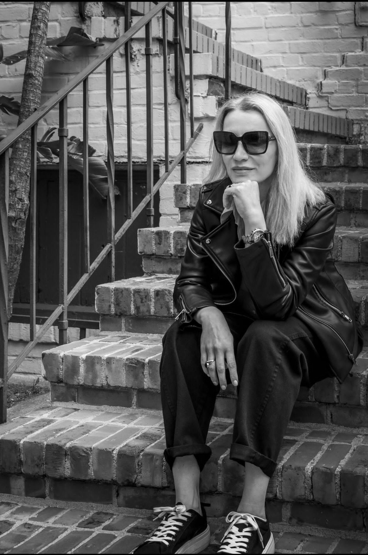 a woman wearing sunglasses and a leather jacket is sitting on a set of stairs .