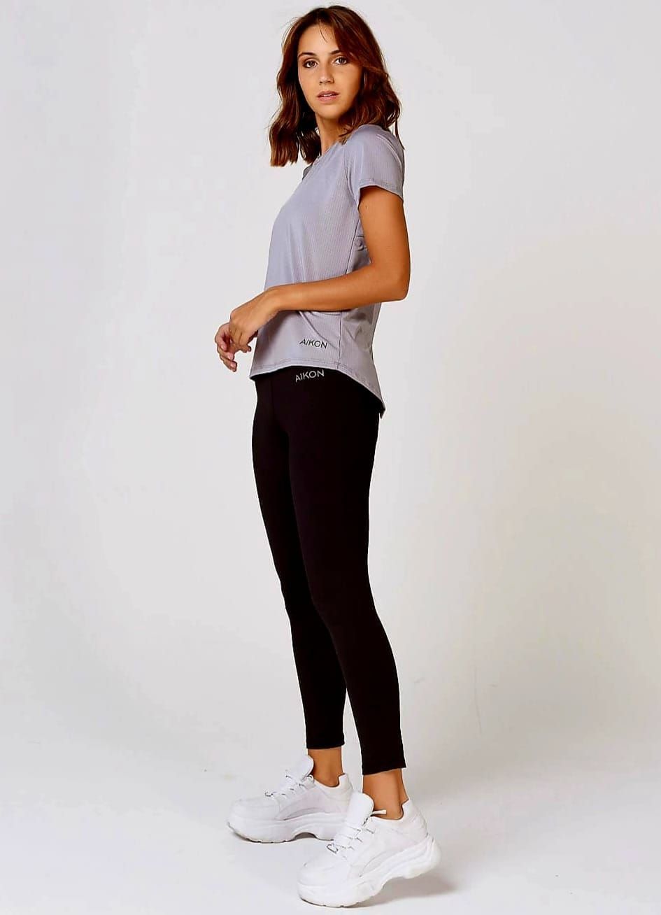 Mujer con top gris y leggings negros, de pie de lado, con zapatillas blancas y contra un fondo blanco.