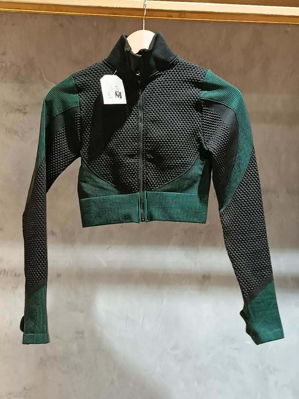 Chaqueta deportiva corta verde y negra con cremallera, colgada de una barra de madera.
