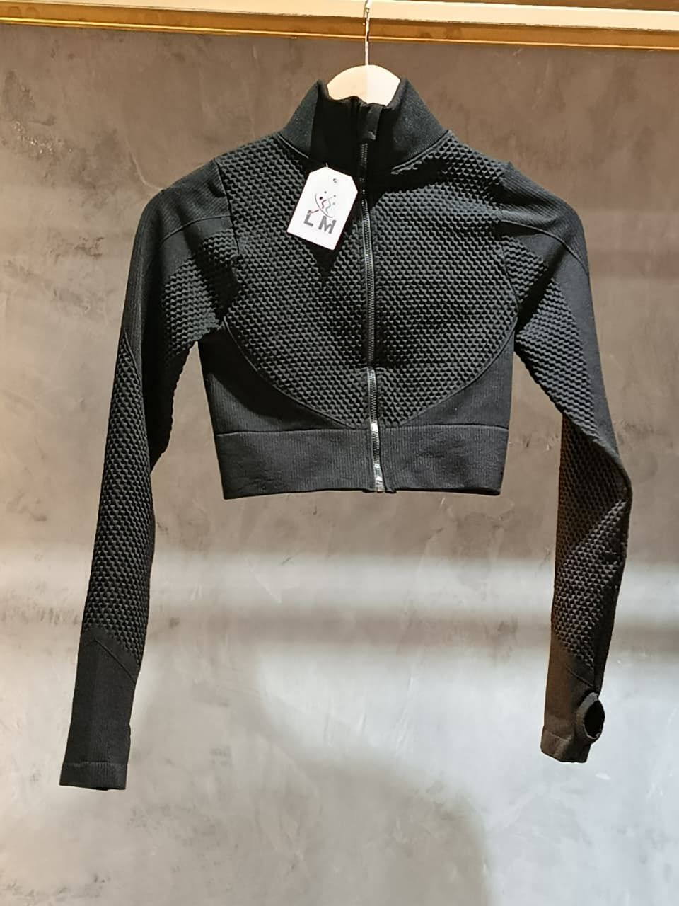 Chaqueta deportiva corta negra con frente texturizado y mangas largas, colgada en un perchero de madera.